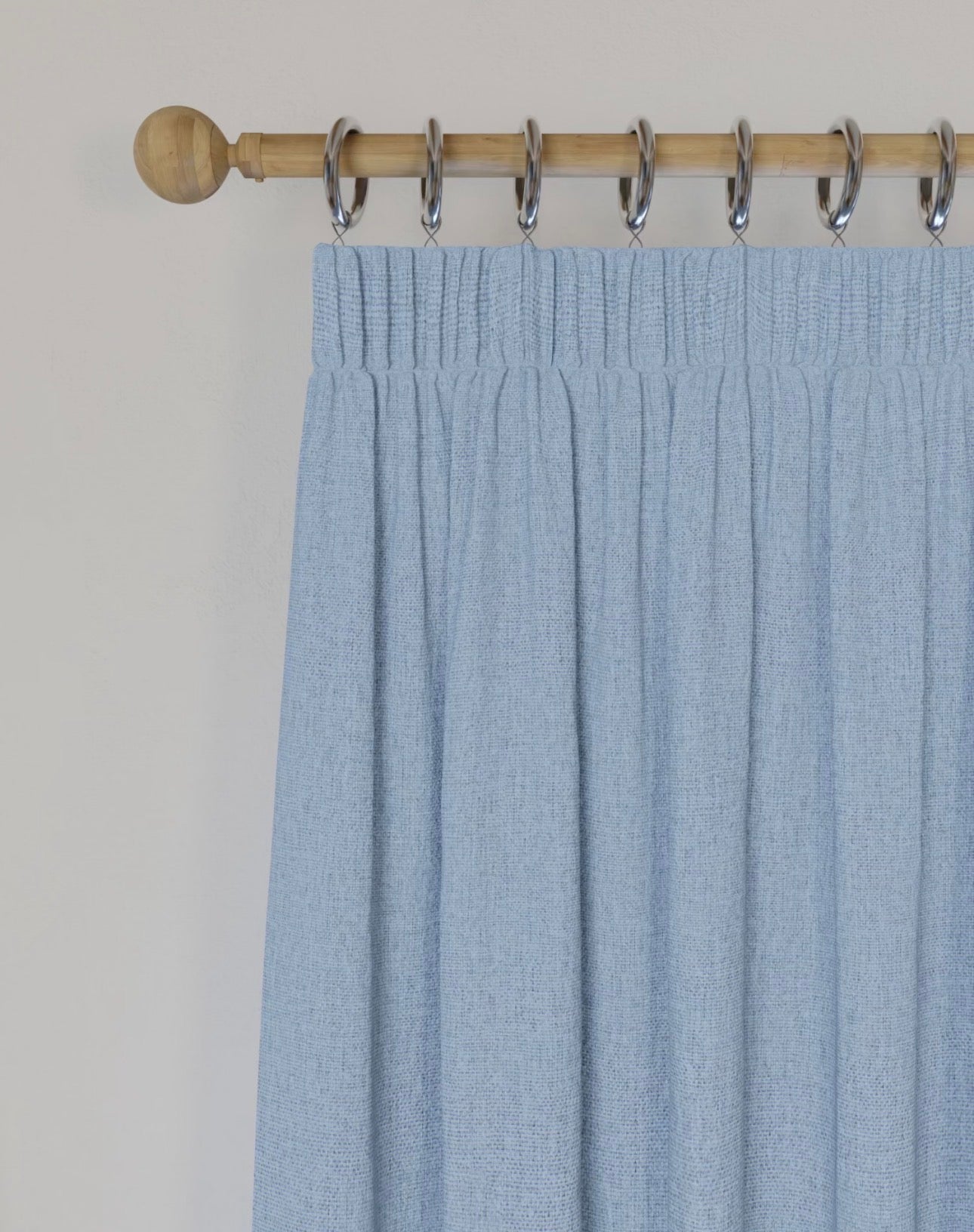 A light blue pleated curtain hanging from a wooden rod with silver metal rings against a plain off-white wall.